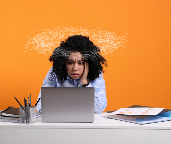 A stressed businesswoman sitting at a desk staring at her laptop with her head resting on her hand, surrounded by paperwork and office supplies, with a scribbled cloud above her head symbolizing burnout against an orange background.