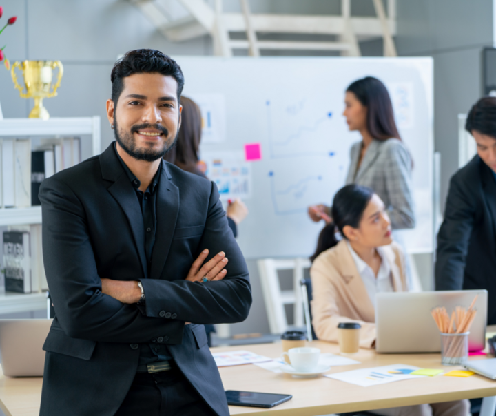 A confident businessman in a black suit stands smiling with arms crossed in a modern office, while colleagues collaborate at a table and whiteboard in the background.